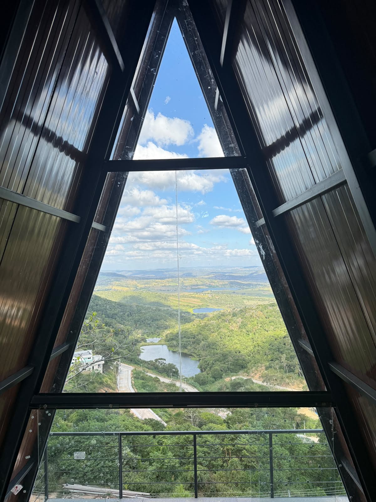 Interior de uma cabana em obra, com a estrutura em A emoldurando a vista do vale e do açude (outubro de 2026)